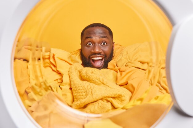 мужчина позирует изнутри стиральной машины или сушилки для белья, собирает желтую одежду для стирки, занятый работой по дому