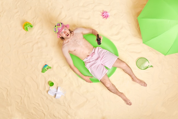 Free photo man poses on swim ring at seaside surrounded by beach accessories drinks beer wears sunhat snorkeling mask and shorts tans in sunlight