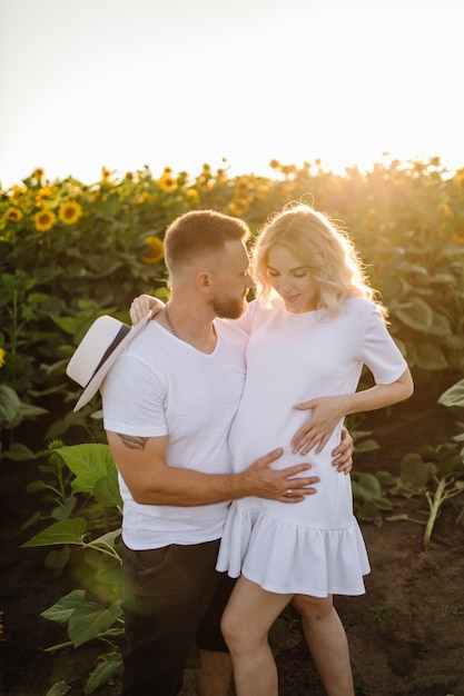 Free photo man and pregnant woman hug each other tender standing in the field with tall sunflowers around them