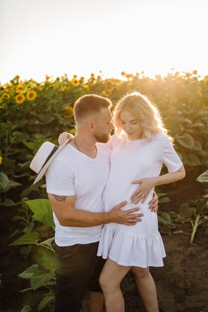 Man and pregnant woman hug each other tender standing in the field with tall sunflowers around them