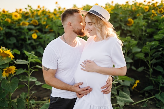 Man and pregnant woman hug each other tender standing in the field with tall sunflowers around them