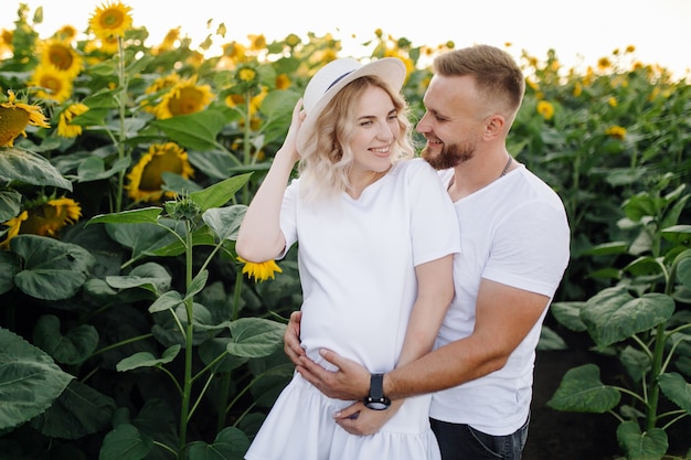 Man and pregnant woman hug each other tender standing in the field with tall sunflowers around them