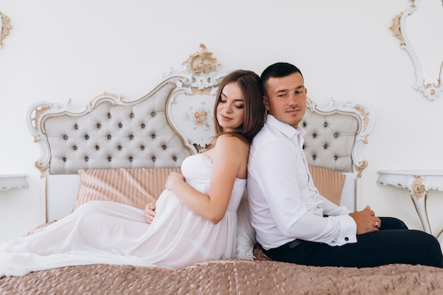 Man and pregnant woman sit side by side on a luxury white bed