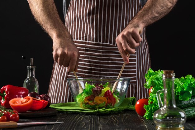 Мужчина готовит салат со свежими овощами на деревянном столе. Приготовление вкусной и здоровой пищи. На черном фоне. Вегетарианская еда, здоровая или кулинарная концепция.