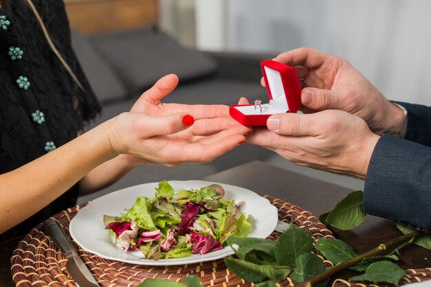 Man presenting gift box with ring to woman at table with plate 