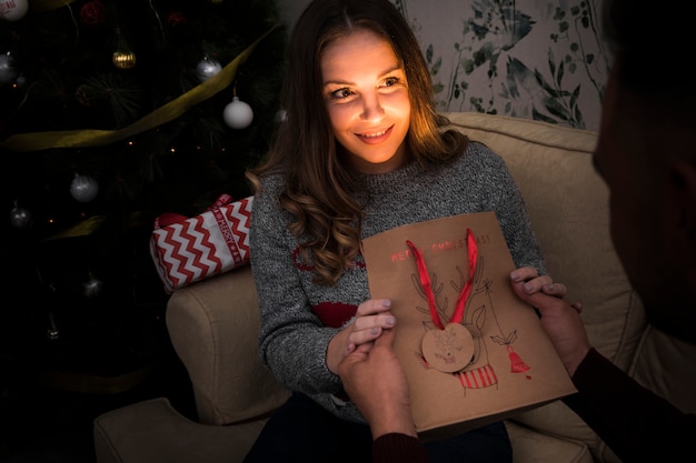 Человек, представляя подарок пакет привлекательной женщине на диване возле Рождественская елка
