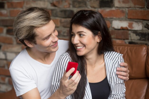 Man proposing to excited woman at home, holding ring box 