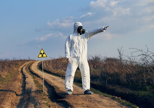 Man in protective coverall and gas mask standing on village road in field next to caution sign