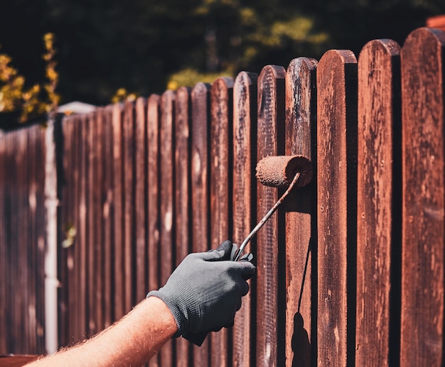 Man in protective gloves is painting wooden fence in bright summer day.