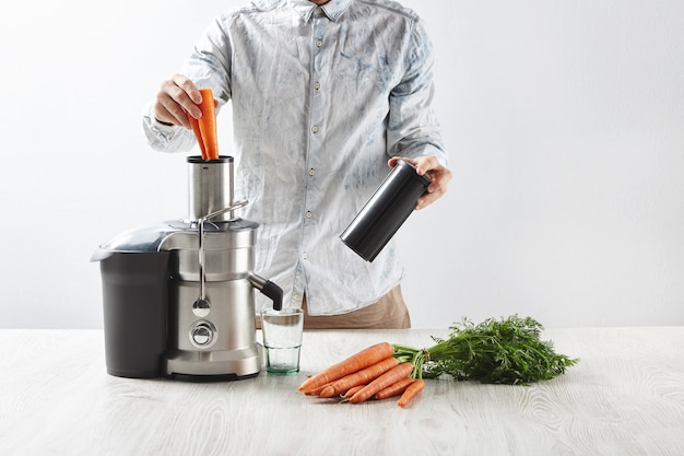 Бесплатное фото Мужчина кладет морковь в металлическую соковыжималку с пустым стаканом, чтобы приготовить вкусный сок на завтрак
