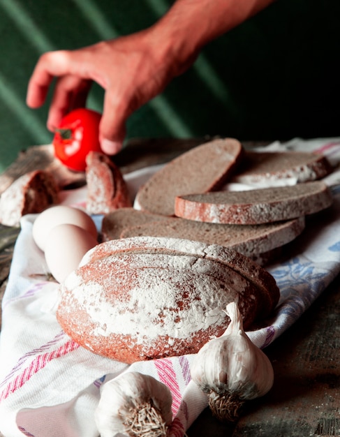 Man putting tomato on a white tablecloth with bread slices, eggs and garlic gloves around.