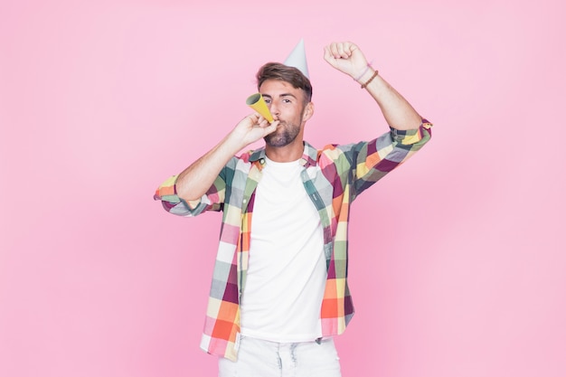 Man raising his arms while blowing party horn on pink background