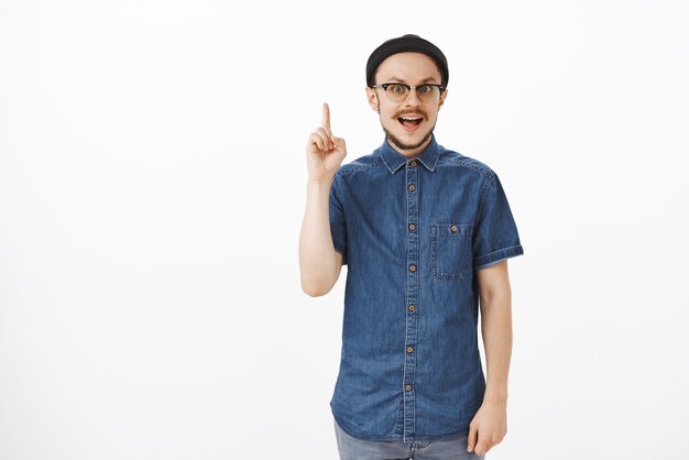 Мужчина поднимает указательный палец в жесте эврики, добавляя предложение сосредоточенным спокойным выражением лица, стоит в черной шапочке и синей рубашке над серой стеной
