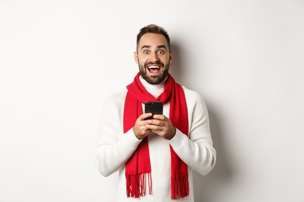 Мужчина читает удивительное промо-предложение в Интернете, держит смартфон и выглядит удивленным, стоя в зимнем свитере на белом фоне.