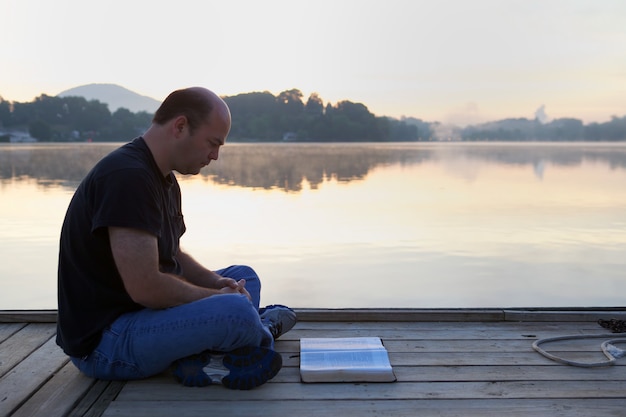 Man reading a book on a wooden bridge surrounded by hills and a lake under the sunlight