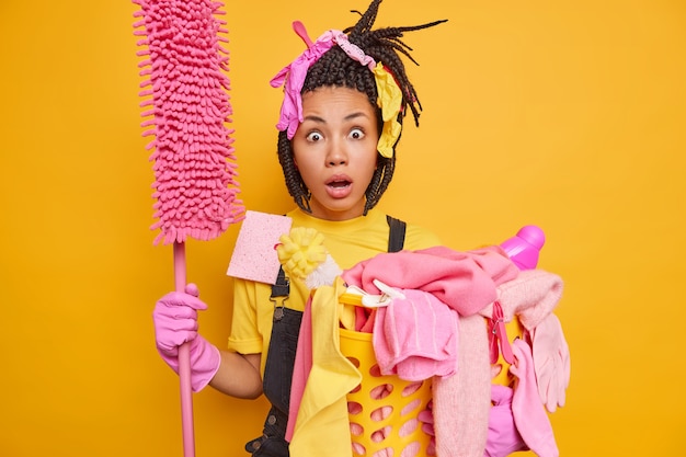 Man ready for cleaning carries mop laundry basket looks with unbelievable expression dressed in onalls rubber gloves isolated on vivid yellow