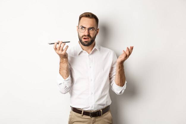 Man recording voice message or talking on speakerphone, looking confused, standing  