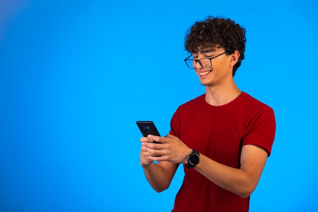 Человек в красной рубашке делает селфи на смартфоне на синем и смеется.