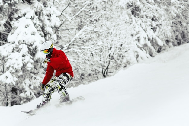 Man in red ski jacket and white helmet goes down the snowed hill in the forest 