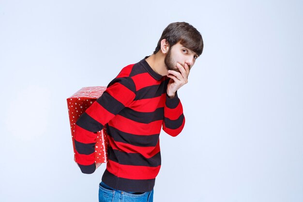 Человек в красной полосатой рубашке прячет за собой красную подарочную коробку.