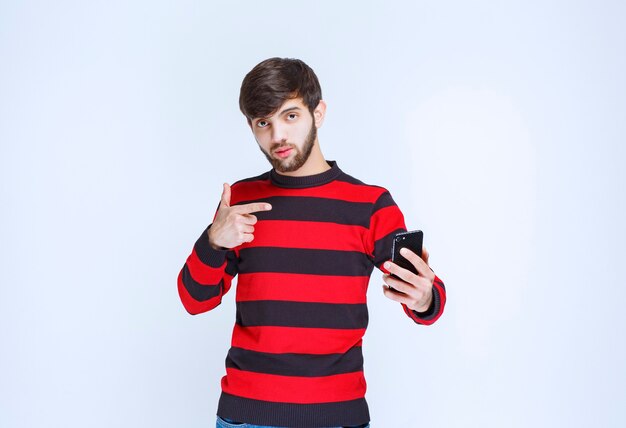 Man in red striped shirt holding a black smartphone and pointing at it.