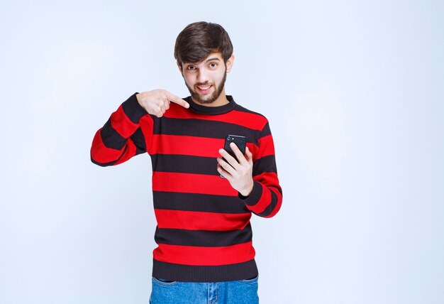 Man in red striped shirt holding a black smartphone and pointing at it.
