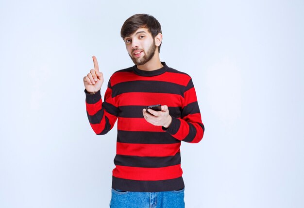 Человек в красной полосатой рубашке держит черный смартфон и думает.