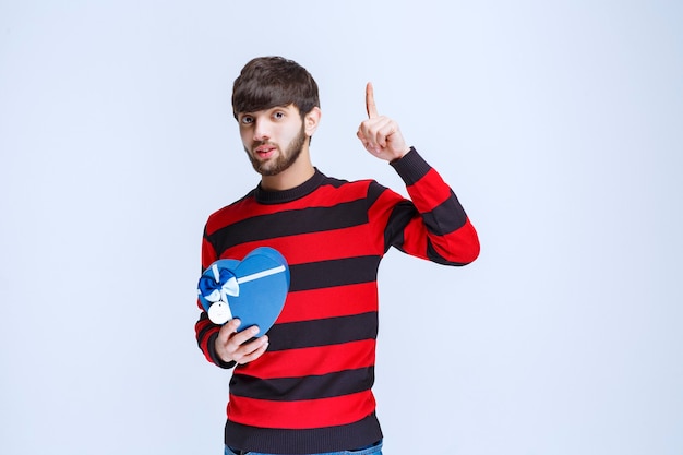 Man in red striped shirt holding a blue heart shape gift box and looks thoughtful or like he has a good idea.