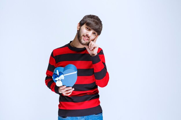Man in red striped shirt holding a blue heart shape gift box and looks thoughtful or like he has a good idea.