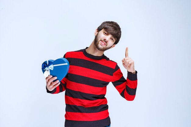 Man in red striped shirt holding a blue heart shape gift box and looks thoughtful or like he has a good idea.