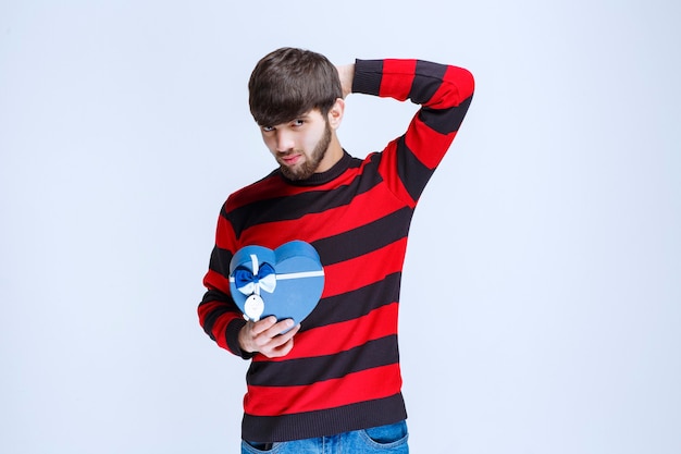 Man in red striped shirt holding a blue heart shape gift box and looks thoughtful or like he has a good idea.