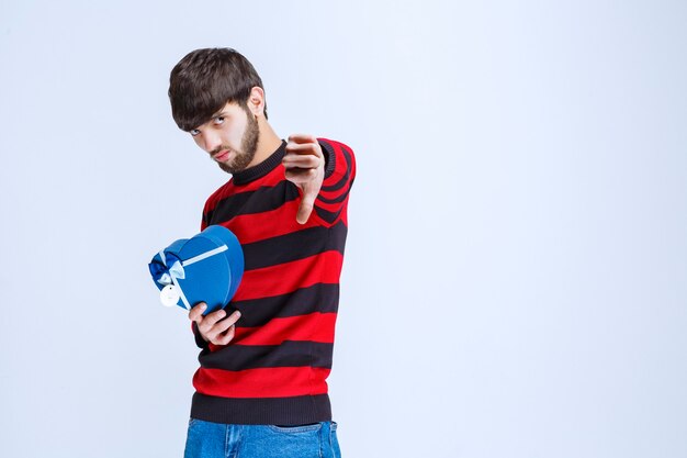 Man in red striped shirt holding a blue heart shape gift box and showing dislike sign.