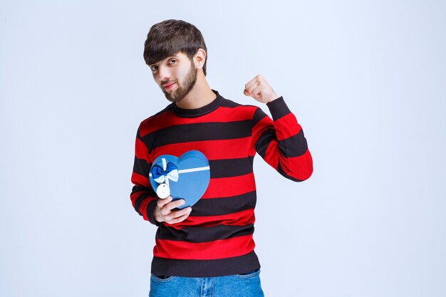 Man in red striped shirt holding a blue heart shape gift box and showing his fist.