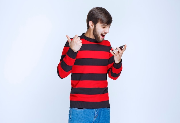 Человек в красной полосатой рубашке держит телефон и делает селфи в энергичных позах.