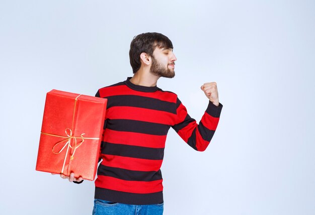 Мужчина в красной полосатой рубашке держит красную подарочную коробку и чувствует себя сильным и позитивным.
