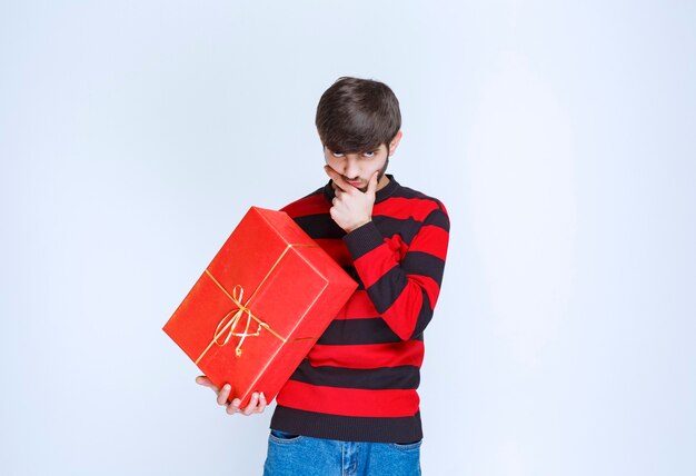 Free photo man in red striped shirt holding a red gift box and looks confused and thoughtful.
