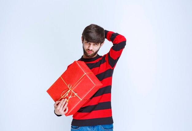 빨간색 선물 상자를 들고 빨간색 줄무늬 셔츠에 남자가 혼란스럽고 사려 깊습니다.