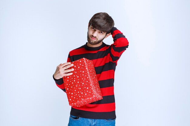 Man in red striped shirt holding a red gift box and looks confused and thoughtful.