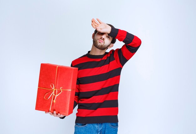 Мужчина в красной полосатой рубашке, держащий красную подарочную коробку, выглядит усталым и сонным.