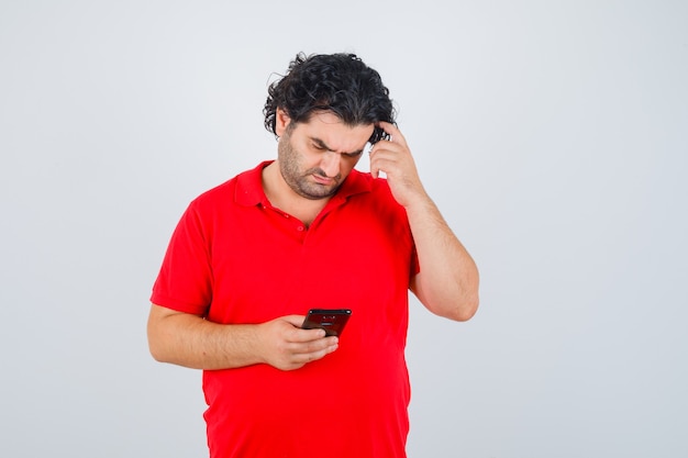 Free photo man in red t-shirt holding phone, scratching head and looking focused