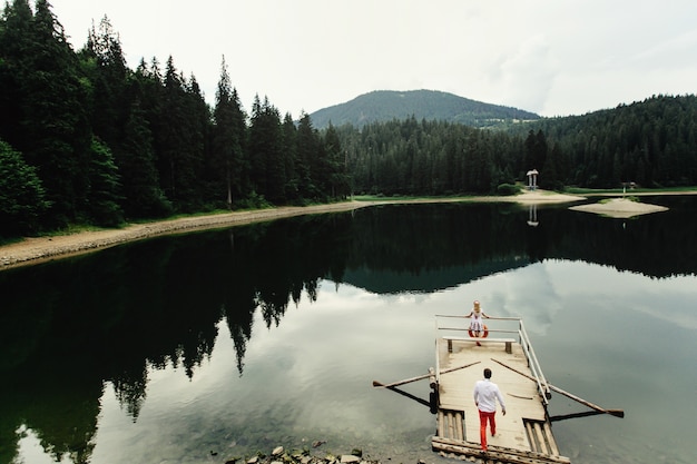 Мужчина в красных брюках ходит к одинокой леди, стоящей на деревянной лодке над горным озером