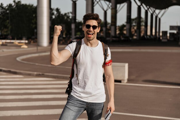 Мужчина радуется и улыбается Красивый брюнет в белой футболке и солнцезащитных очках держит рюкзак и паспорт возле аэропорта