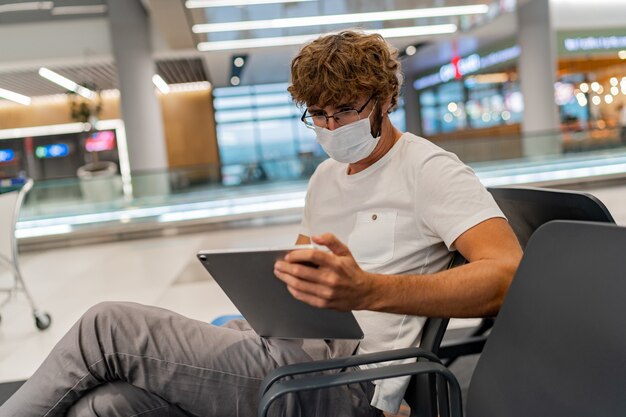 인공 호흡기 마스크를 쓴 남자가 공항에서 다음 비행기를 기다리고 태블릿을 사용하고 있습니다.