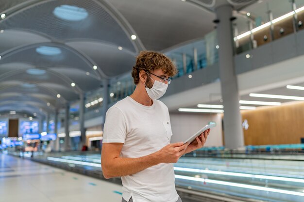 レスピレーターマスクの男が空港で次の飛行機を待っており、タブレットを使用しています。