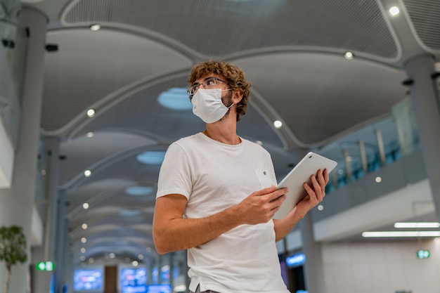 レスピレーターマスクの男が空港で次の飛行機を待っており、タブレットを使用しています。