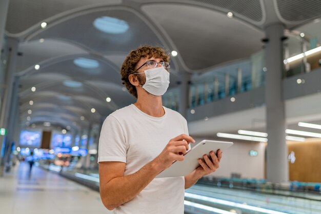 レスピレーターマスクの男が空港で次の飛行機を待っており、タブレットを使用しています。