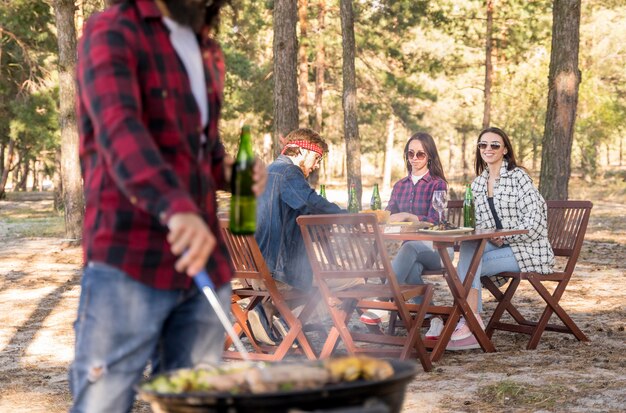 Man roasting corn on barbecue while friends converse at table outdoors