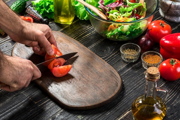 Man's hand cuts ripe red tomatoes for summer healthy vegetable salad on a wooden board, arranged with cucumber, lettuce, parsley, vertical. Top view. Still life. Flat lay Copy space
