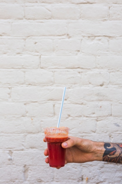 A man's hand holding smoothie in plastic disposable cup with straw against wall