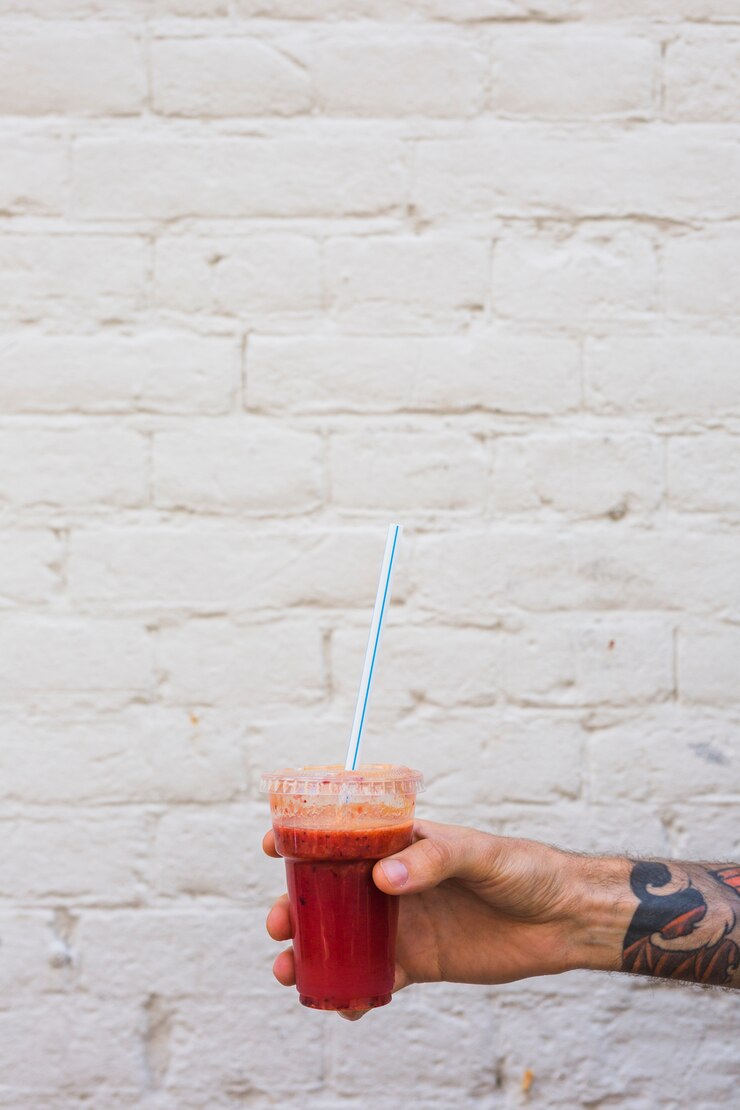 Free Photo a man's hand holding smoothie in plastic disposable cup with straw against wall